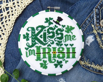 Könnte beinhalten: Ein weißer Stickrahmen mit grünen und weißen Stichen. Der Rahmen zeigt den Text "Kiss me, I'm Irish" mit einem Kleeblatt und einem Koboldhut.