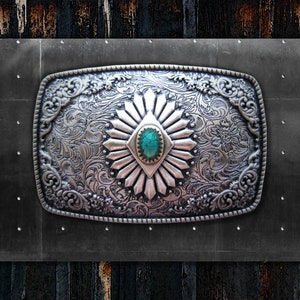 May include: Silver-colored rectangular belt buckle with intricate floral and scrollwork designs. The buckle features a central turquoise-colored stone and a sunburst pattern. The background is a dark gray metal panel with rivets.
