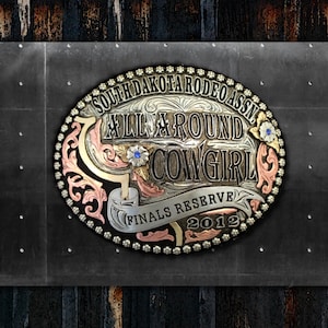 Op de afbeelding: Een zilveren, gouden en koperen gesp met een bloemmotief en de tekst "South Dakota Rodeo Assn. All Around Cowgirl Finals Reserve 2012".