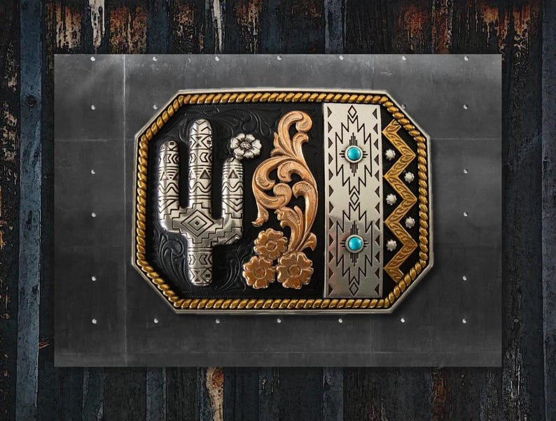 Puede incluir: Hebilla de cintur&oacute;n decorativa con un cactus plateado, detalles florales dorados y piedras turquesas. La hebilla tiene un fondo negro y un borde plateado y dorado. El dise&ntilde;o est&aacute; sobre un fondo gris oscuro.