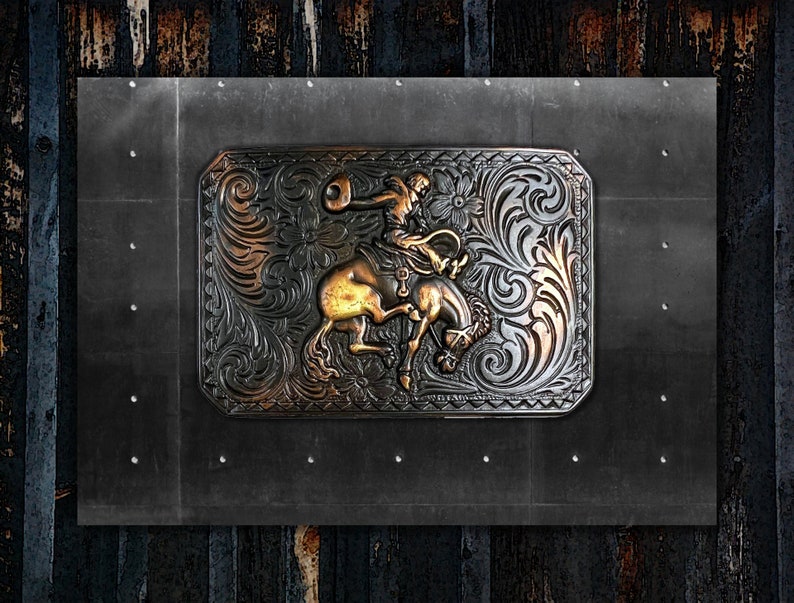 Puede incluir: Una hebilla de cintur&oacute;n de metal plateado y dorado con un dise&ntilde;o intrincado de un vaquero montando un caballo. La hebilla tiene un patr&oacute;n floral y un dise&ntilde;o en relieve.