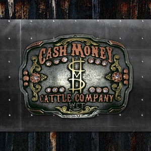 May include: A silver belt buckle with a green background and gold accents. The buckle features the text "CASH MONEY CATTLE COMPANY WCT" and a stylized dollar sign with a cross through it.