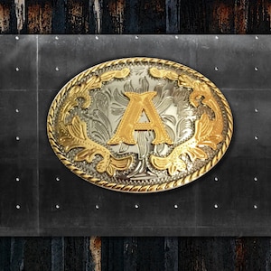 May include: A silver and gold belt buckle with an ornate design and the letter "A" in gold in the center.