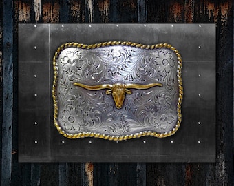 Puede incluir: Una hebilla de cinturón de plata con un intrincado diseño floral y una cabeza de toro longhorn dorada en el centro. La hebilla está rodeada por un borde de cuerda dorada.