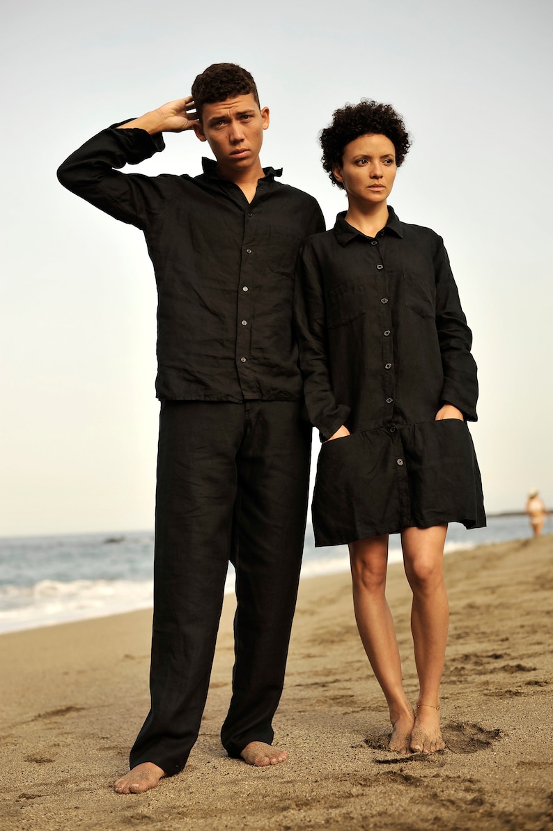 Pu&ograve; includere: Due persone con camicie di lino nere in piedi su una spiaggia di sabbia. L'uomo indossa una camicia a maniche lunghe e pantaloni. La donna indossa un abito camicia a maniche lunghe.