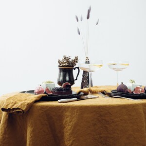 May include: A table setting with a brown linen tablecloth, two champagne flutes, a black and gold ceramic mug, a black plate with a dessert and figs, and a white knife.