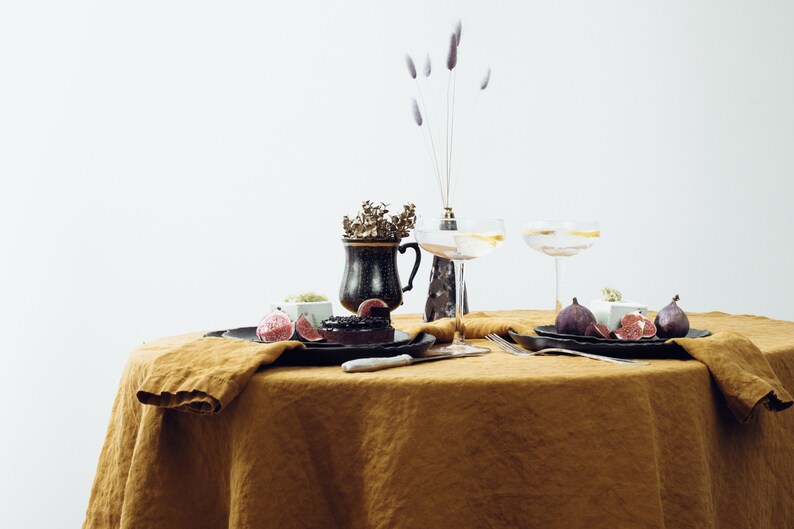 May include: A table setting with a brown linen tablecloth, two champagne flutes, a black and gold ceramic mug, a black plate with a dessert and figs, and a white knife.