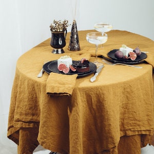 May include: A round, mustard yellow linen tablecloth with two place settings. Each place setting has a black plate, a silver fork and knife, and a white napkin. A small dessert with a white topping and a dark chocolate base is on one plate. A small vase with dried flowers and a larger vase with dried flowers are on the table.