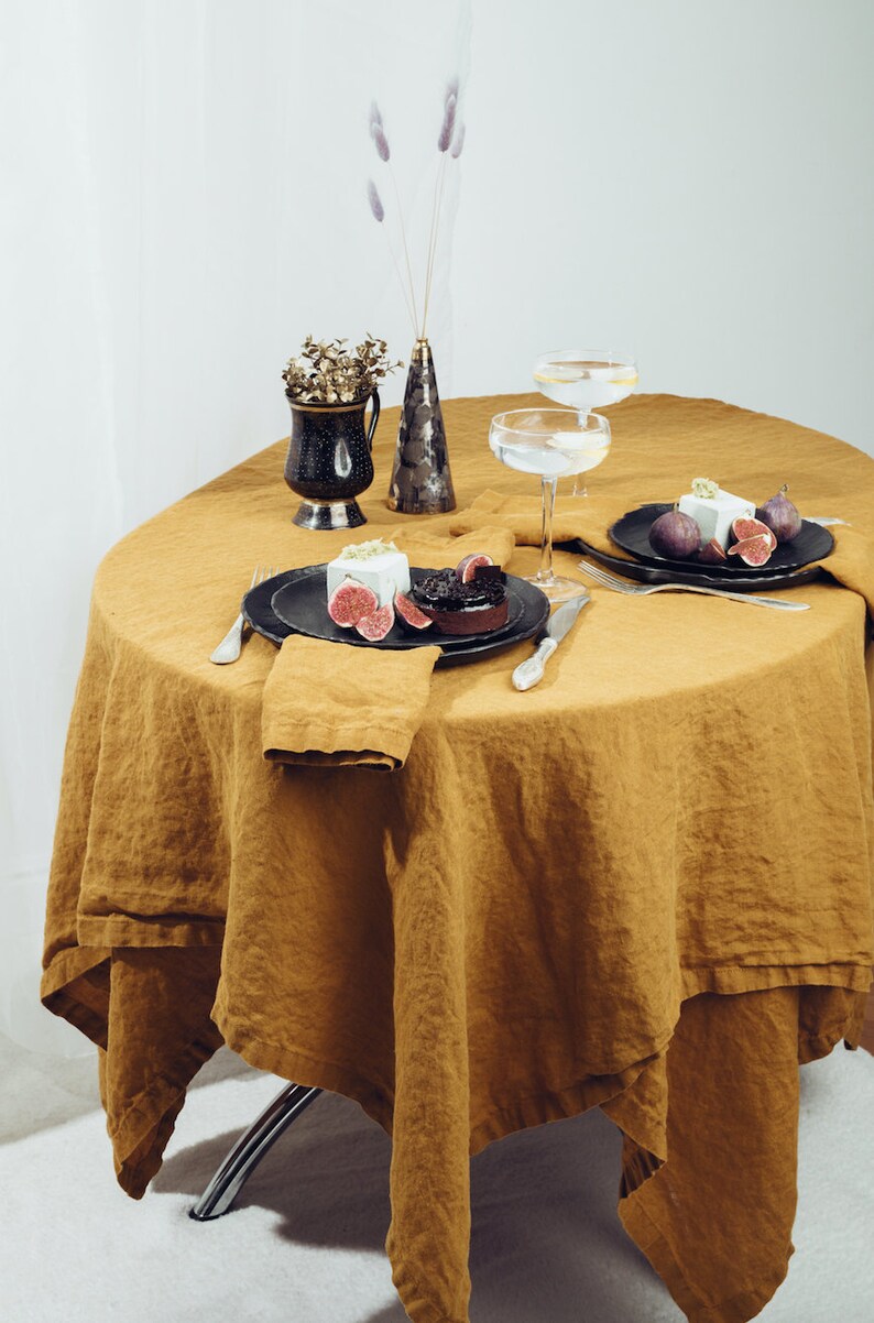 May include: A round, mustard yellow linen tablecloth with two place settings. Each place setting has a black plate, a silver fork and knife, and a white napkin. A small dessert with a white topping and a dark chocolate base is on one plate. A small vase with dried flowers and a larger vase with dried flowers are on the table.