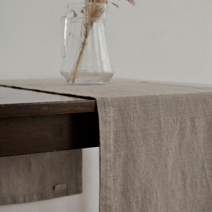 Pode incluir: Um corredor de mesa de linho castanho claro sobre uma mesa de madeira castanho escuro. Um jarro de vidro transparente com erva seca está sobre a mesa.