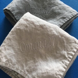 K&ouml;nnte beinhalten: Zwei Leinenservietten mit dem gestickten Text "Omama27". Die obere Serviette ist grau und die untere Serviette ist beige.
