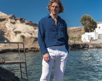 Può includere: Un uomo con una camicia di lino blu scuro e pantaloni di lino bianchi si trova in piedi su una costa rocciosa. L'acqua è blu e il cielo è sereno.