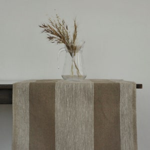 May include: A brown and beige striped table runner with a clear glass pitcher holding dried wheat stalks. The runner is draped over a dark wooden table, creating a neutral, rustic aesthetic.