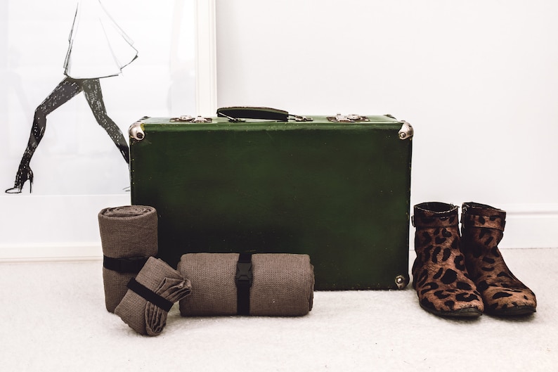May include: An olive green vintage suitcase with silver corner protectors and a black handle. Rolled brown blankets are in front of the suitcase, secured with black straps. Leopard print ankle boots are to the right, and a framed fashion illustration is in the background.