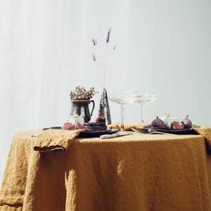 May include: A table setting with a brown linen tablecloth, two champagne flutes, a small vase with dried flowers, and two plates with figs and a dessert.