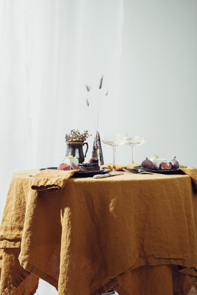 May include: A table setting with a brown linen tablecloth, two champagne flutes, a small vase with dried flowers, and two plates with figs and a dessert.