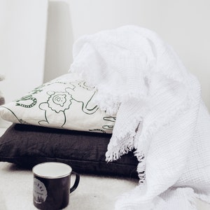 May include: A white throw blanket with a fringed edge is draped over two cushions, one with a green and white animal print and the other a dark grey linen. A black enamel mug with a white design is on the floor in front of the cushions.