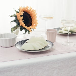 May include: A white tablecloth with a pink linen runner. A sunflower in a glass vase, a plate of melon slices, two glasses of white wine, and two small cups are on the table.