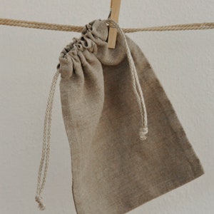 May include: A beige linen drawstring bag hanging from a rope with a wooden clothespin. The bag is empty and has a natural, rustic look.