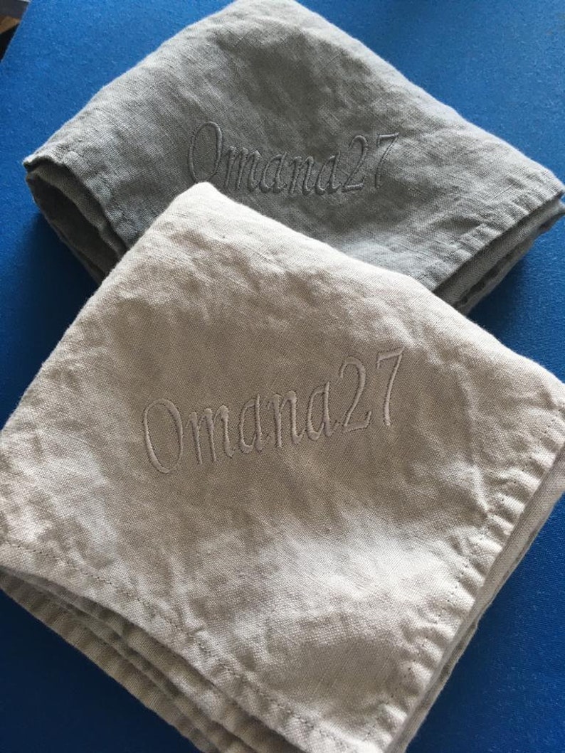 以下が含まれることがあります： 「Omama27」と刺繍されたリネン製のナプキン2枚。上のナプキンはグレーで、下のナプキンはベージュです。