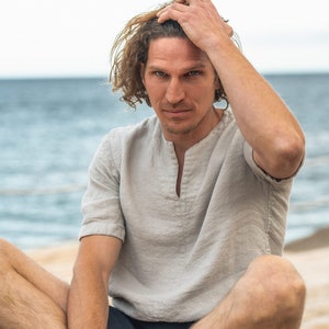 May include: A man with long, wavy brown hair sits with his legs crossed, wearing a light grey, short-sleeved shirt and dark blue shorts. He is sitting on a wooden surface with the sea in the background.
