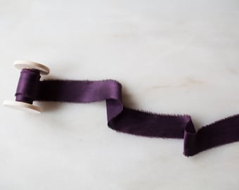 May include: A spool of deep purple silk ribbon with frayed edges. The ribbon is wound around a wooden spool.