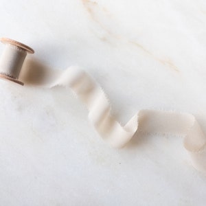 Puede incluir: Una bobina de cinta de seda de color crema con una bobina de madera. La cinta está sobre una superficie de mármol blanco.