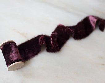 May include: A spool of burgundy velvet ribbon with a wooden spool. The ribbon is curled and draped on a white marble surface.