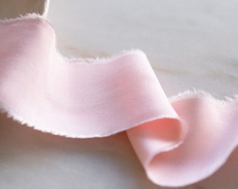May include: A pale pink ribbon with a raw edge, folded and curled on a light wood surface.
