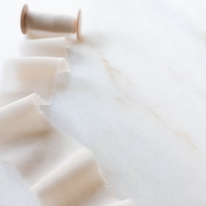 May include: A spool of off-white silk ribbon with a wooden spool and a long length of ribbon unfurling on a white marble surface.