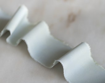 May include: A light blue satin ribbon with a raw edge, curled and folded on a white surface.