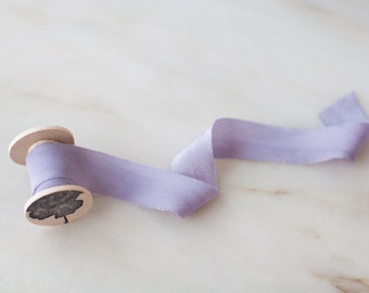 May include: A spool of lavender silk ribbon with a wooden spool and a decorative design on the end.
