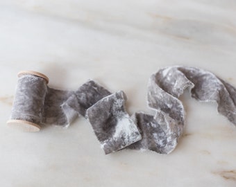 May include: A spool of gray velvet ribbon with a wooden spool. The ribbon is unraveled and has a slightly textured look.