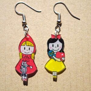 Peut inclure: Une paire de boucles d'oreilles représentant des personnages de dessins animés. La boucle d'oreille gauche représente une fille à capuchon rouge tenant un loup. La boucle d'oreille droite représente une fille aux cheveux noirs tenant une pomme rouge.