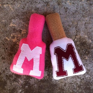May include: Two plush fabric letters, both shaped like the letter "M". One is pink with a white "M" and the other is white with a maroon "M".