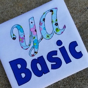 Puede incluir: Camiseta blanca con el texto "ya basic" en azul y bordado floral. La palabra "ya" está en una fuente cursiva y la palabra "basic" está en una fuente en negrita.