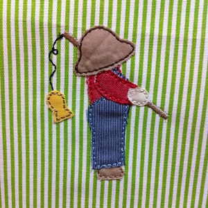 May include: A green and white striped fabric with an embroidered image of a person wearing a brown hat, red shirt, blue overalls, and holding a fishing rod. The person is standing on a green and white striped background.
