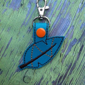 Puede incluir: Un llavero de cuero azul turquesa en forma de hoja con una vena cosida marrón y un botón naranja. El llavero tiene un cierre de metal plateado.