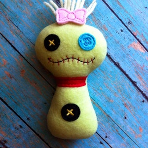 以下が含まれることがあります： 黄色い体、黒いボタンの目、片方の目に青いボタン、頭にピンクのリボンが付いた手作りのかやの人形。人形は青と白の木製の表面の上に座っています。