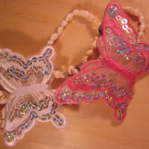 May include: Two butterfly-shaped ornaments, one white and one pink, are adorned with iridescent sequins. They are connected to a necklace made of small seashells. The ornaments are set against a light brown wooden surface.