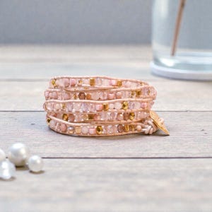 May include: A multi-strand wrap bracelet with pink and gold beads. The bracelet is wrapped around multiple times and fastened with a leather strap and a gold-coloured charm. The beads are various shapes and sizes, creating a delicate accessory.