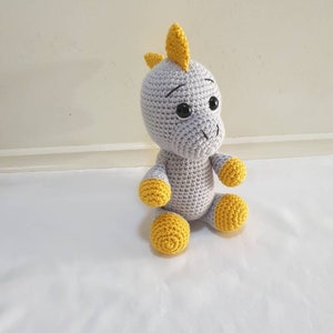 May include: A gray crocheted dinosaur toy with yellow accents. The dinosaur has large black eyes and a yellow horn on its head.