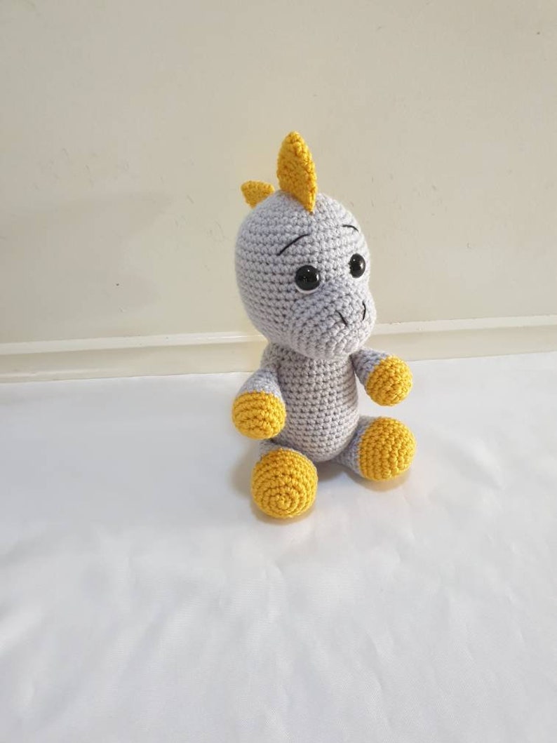 May include: A gray crocheted dinosaur toy with yellow accents. The dinosaur has large black eyes and a yellow horn on its head.