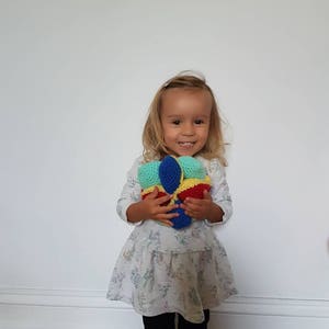 May include: A young girl in a white dress with a floral pattern holds a colorful crocheted ball. The ball is made of blue, yellow, red, and green yarn.
