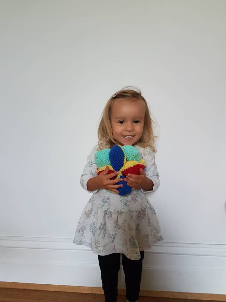 May include: A young girl in a white dress with a floral pattern holds a colorful crocheted ball. The ball is made of blue, yellow, red, and green yarn.