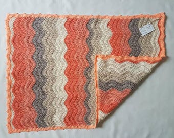 May include: A crocheted baby blanket with a wavy pattern in shades of peach, beige, and gray. The blanket has a scalloped edge.