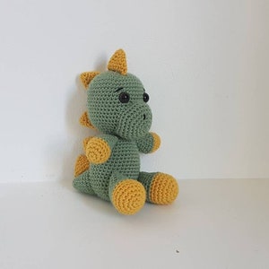 May include: A green crocheted dinosaur toy with yellow accents. The dinosaur has a small, pointed nose and large, black eyes.