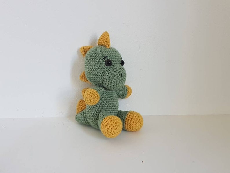 May include: A green crocheted dinosaur toy with yellow accents. The dinosaur has a small, pointed nose and large, black eyes.