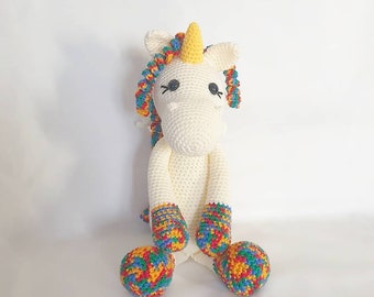 May include: A white crocheted unicorn with a yellow horn and rainbow colored mane and hooves. The unicorn is sitting with its legs crossed.