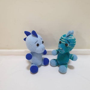 May include: Two crocheted stuffed animals, one blue and one teal, sitting on a white surface. Both animals have blue eyes and a blue nose.
